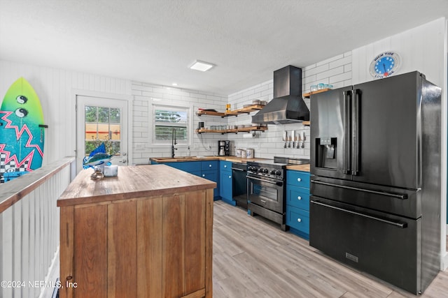 kitchen featuring black appliances, blue cabinetry, sink, wall chimney range hood, and light wood-type flooring