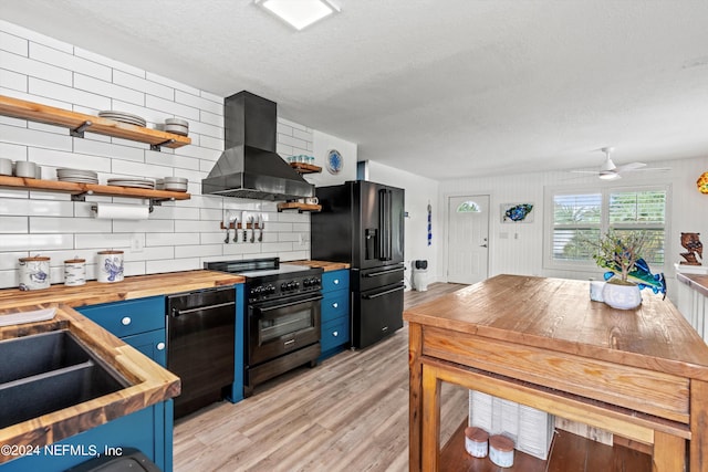 kitchen featuring blue cabinetry, butcher block countertops, black appliances, wall chimney range hood, and light hardwood / wood-style floors