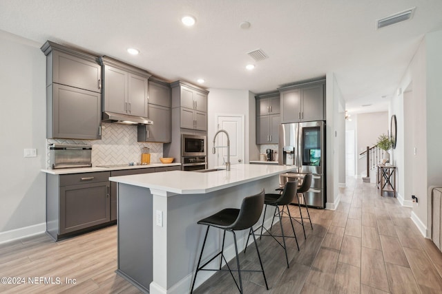 kitchen featuring light wood-type flooring, appliances with stainless steel finishes, gray cabinets, and an island with sink