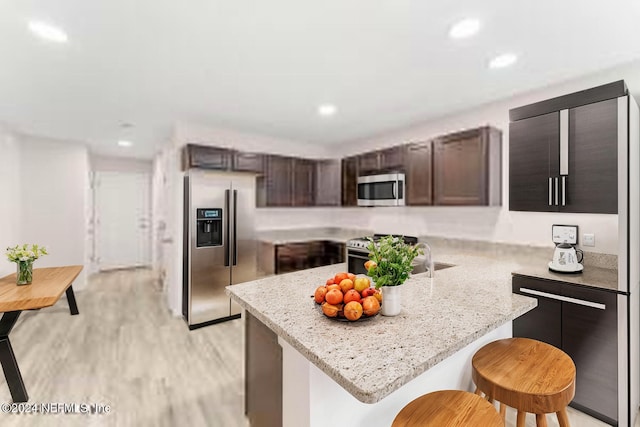 kitchen with dark brown cabinetry, appliances with stainless steel finishes, a breakfast bar, light stone countertops, and light hardwood / wood-style floors