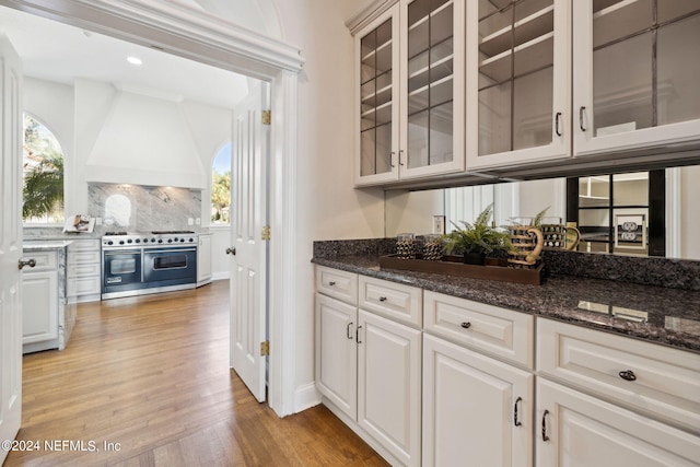 kitchen with white cabinets, premium range hood, light hardwood / wood-style flooring, dark stone countertops, and range with two ovens