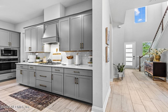 kitchen with backsplash, appliances with stainless steel finishes, custom range hood, gray cabinetry, and light hardwood / wood-style floors