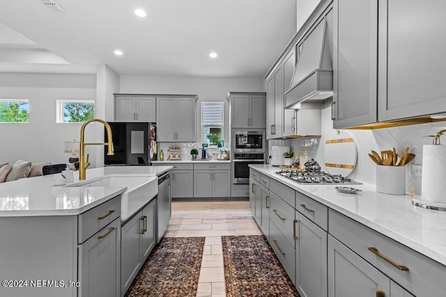 kitchen featuring gray cabinets, a kitchen island with sink, custom exhaust hood, stainless steel appliances, and decorative backsplash