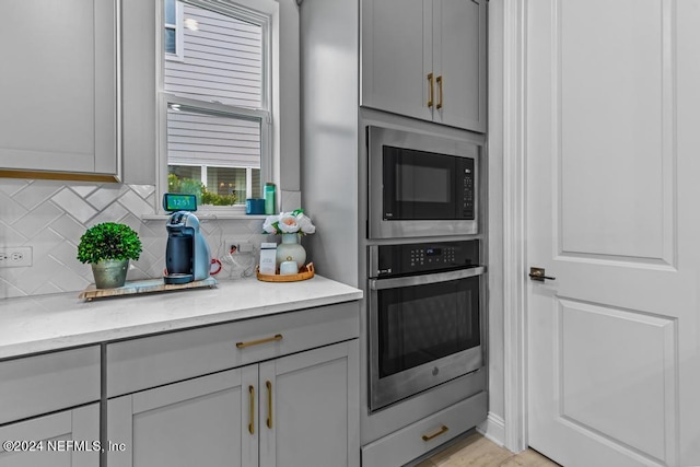 kitchen featuring gray cabinets, backsplash, light stone counters, stainless steel oven, and black microwave