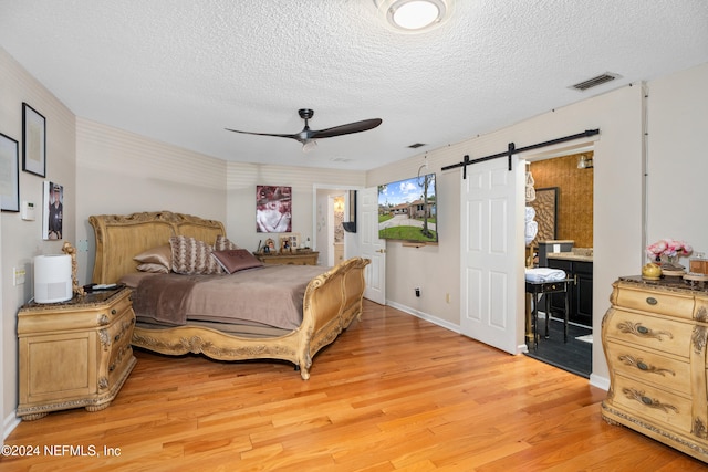 bedroom featuring ensuite bathroom, light hardwood / wood-style floors, a textured ceiling, a barn door, and ceiling fan