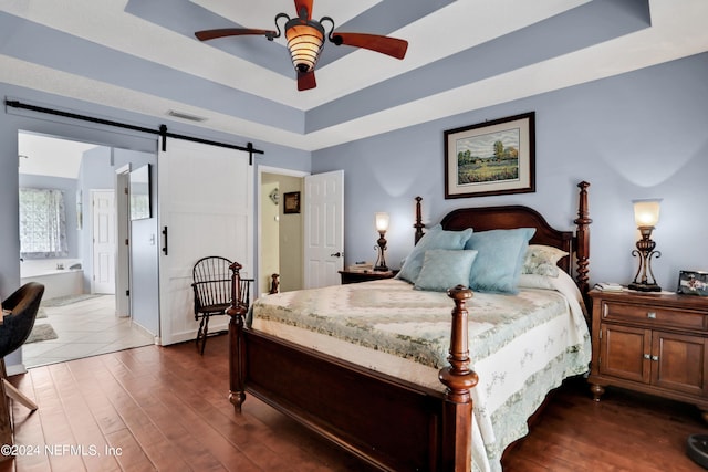 bedroom with ensuite bath, a barn door, dark hardwood / wood-style flooring, a raised ceiling, and ceiling fan