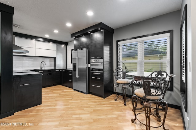 kitchen with a textured ceiling, light hardwood / wood-style flooring, decorative backsplash, stainless steel fridge with ice dispenser, and wall chimney exhaust hood