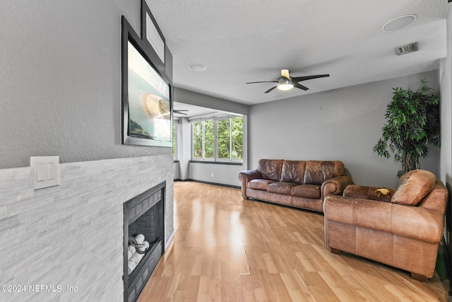 living room featuring light wood-type flooring, a textured ceiling, ceiling fan, and a stone fireplace