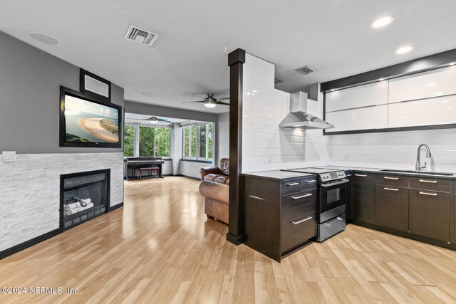 kitchen featuring a fireplace, stainless steel electric range oven, ceiling fan, wall chimney range hood, and light wood-type flooring