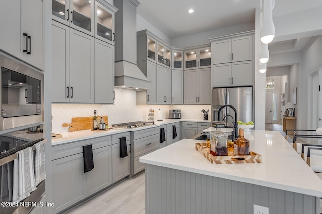 kitchen featuring a kitchen island with sink, appliances with stainless steel finishes, premium range hood, and hanging light fixtures