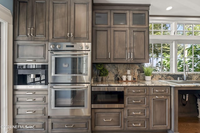 kitchen with light stone countertops, stainless steel double oven, dark brown cabinetry, and sink