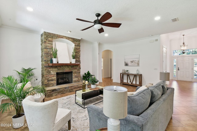 tiled living room featuring ceiling fan with notable chandelier, a stone fireplace, a textured ceiling, and crown molding