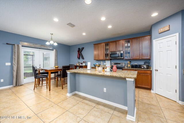 kitchen with decorative light fixtures, appliances with stainless steel finishes, light stone counters, an island with sink, and a textured ceiling