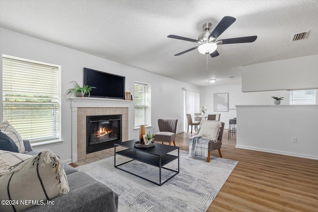 living room featuring a tiled fireplace, ceiling fan, light hardwood / wood-style floors, and a textured ceiling