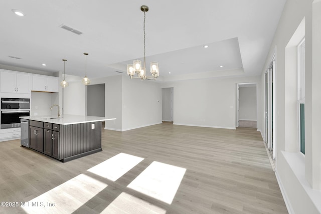 kitchen featuring white cabinetry, hanging light fixtures, light wood-type flooring, a raised ceiling, and an island with sink