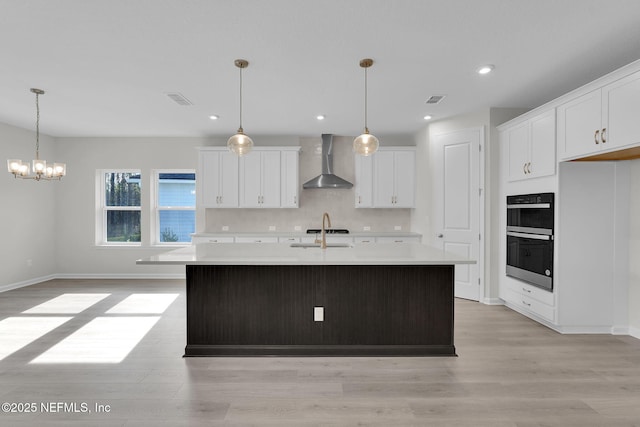 kitchen with a center island with sink, black double oven, wall chimney range hood, and white cabinets