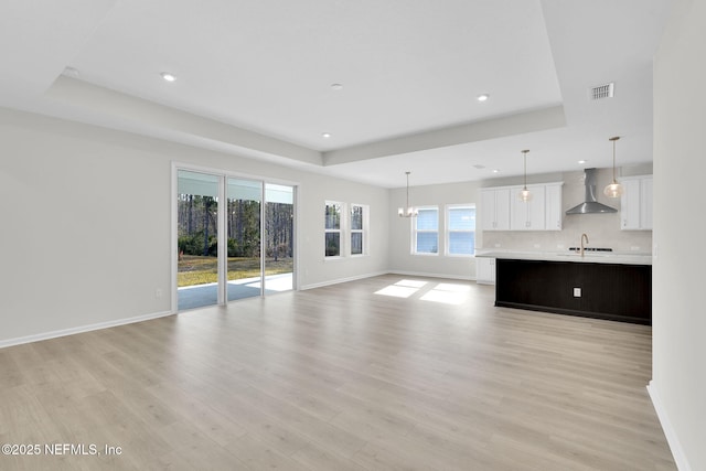 unfurnished living room with sink, a tray ceiling, a notable chandelier, and light wood-type flooring