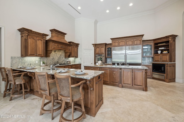 kitchen with kitchen peninsula, a center island with sink, ornamental molding, built in appliances, and a towering ceiling