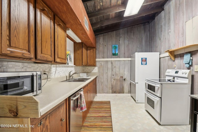 kitchen with wooden walls, stainless steel appliances, a sink, brown cabinetry, and beamed ceiling