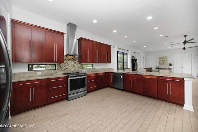 kitchen with light stone counters, kitchen peninsula, wall chimney exhaust hood, appliances with stainless steel finishes, and ceiling fan