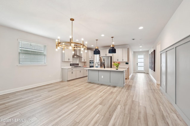 kitchen with light wood-style flooring, light countertops, appliances with stainless steel finishes, wall chimney exhaust hood, and an island with sink