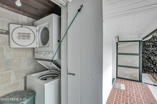 laundry room with wooden ceiling, brick floor, stacked washer and dryer, laundry area, and concrete block wall