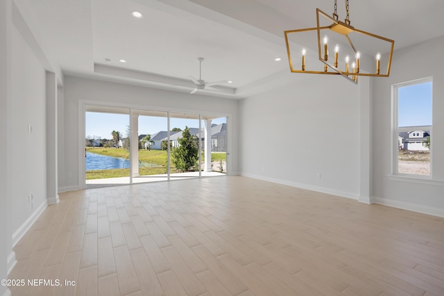 spare room with a raised ceiling, ceiling fan with notable chandelier, a water view, and light wood-type flooring