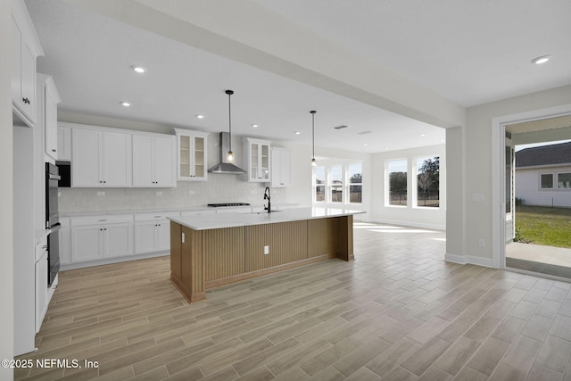 kitchen with decorative light fixtures, tasteful backsplash, an island with sink, white cabinets, and wall chimney range hood