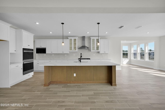 kitchen featuring pendant lighting, double oven, an island with sink, white cabinets, and wall chimney exhaust hood