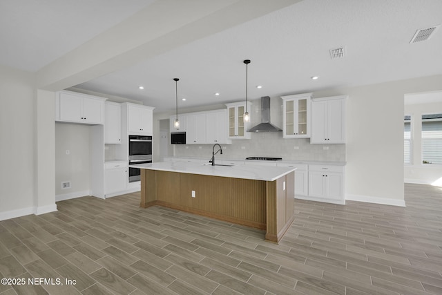 kitchen featuring white cabinetry, wall chimney range hood, decorative light fixtures, and a large island with sink