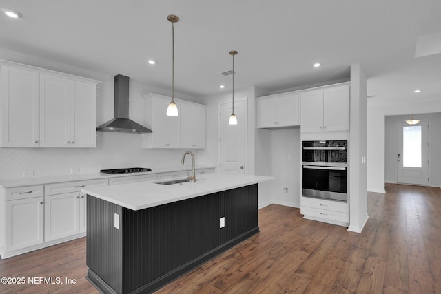 kitchen with an island with sink, stainless steel appliances, wall chimney range hood, white cabinetry, and a sink