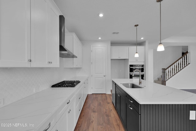 kitchen featuring a center island with sink, white cabinets, wall chimney exhaust hood, hanging light fixtures, and a sink