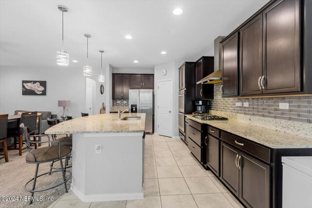 kitchen with hanging light fixtures, a center island with sink, wall chimney range hood, and light stone counters