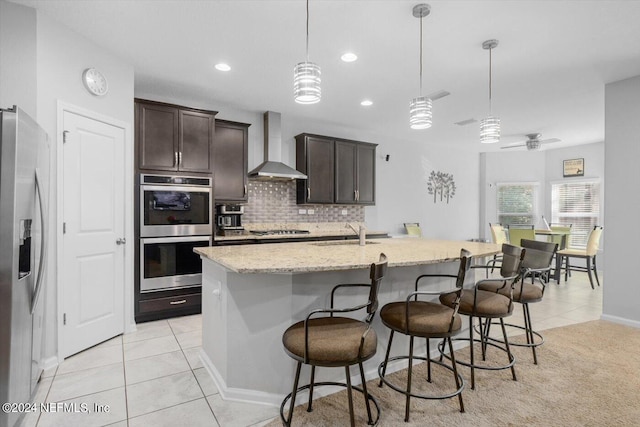 kitchen with ceiling fan, an island with sink, decorative light fixtures, wall chimney exhaust hood, and appliances with stainless steel finishes