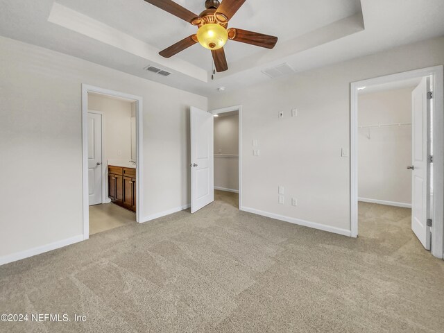 unfurnished bedroom with light carpet, a raised ceiling, a walk in closet, ensuite bathroom, and ceiling fan