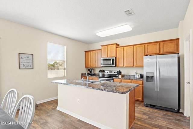 kitchen with appliances with stainless steel finishes, sink, dark stone counters, a kitchen island with sink, and dark wood-type flooring