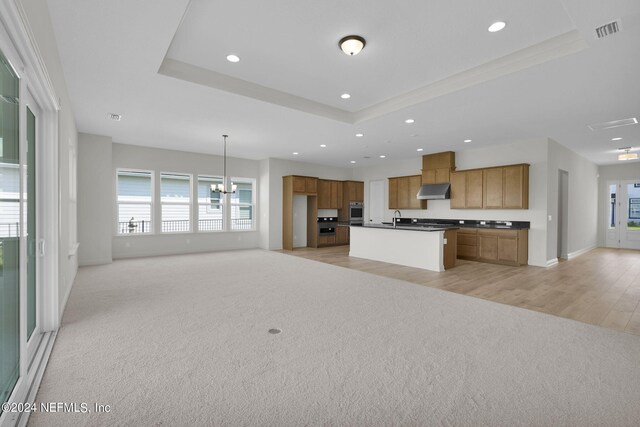 kitchen with a raised ceiling, a center island with sink, and light colored carpet