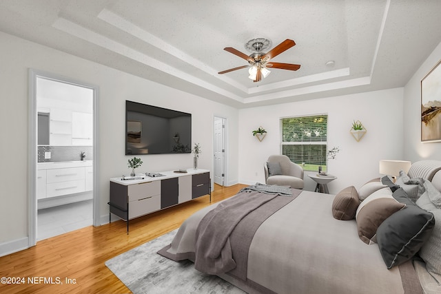 bedroom with light wood-type flooring, baseboards, connected bathroom, a tray ceiling, and ceiling fan