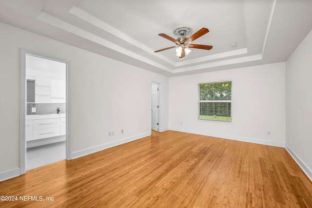 unfurnished bedroom with a tray ceiling, baseboards, ensuite bath, and light wood-style floors