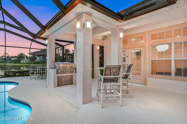 patio terrace at dusk featuring a lanai, exterior bar, a water view, an outdoor kitchen, and a grill