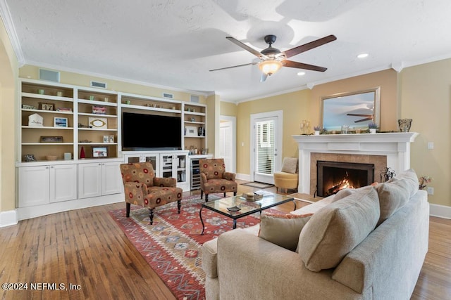 living room featuring ceiling fan, built in shelves, ornamental molding, light hardwood / wood-style floors, and a tiled fireplace