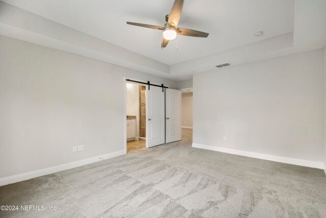 unfurnished bedroom featuring light carpet, ceiling fan, ensuite bathroom, and a barn door