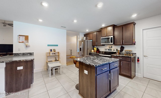 kitchen featuring light tile patterned floors, dark brown cabinets, stainless steel appliances, a center island, and dark stone countertops