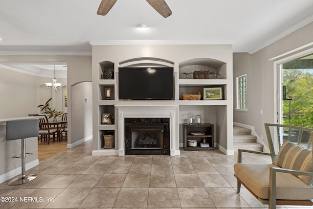 tiled living room featuring a premium fireplace, ornamental molding, ceiling fan with notable chandelier, and built in shelves