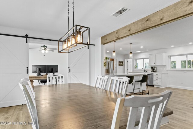 dining area featuring light hardwood / wood-style floors, a barn door, ceiling fan with notable chandelier, beam ceiling, and sink