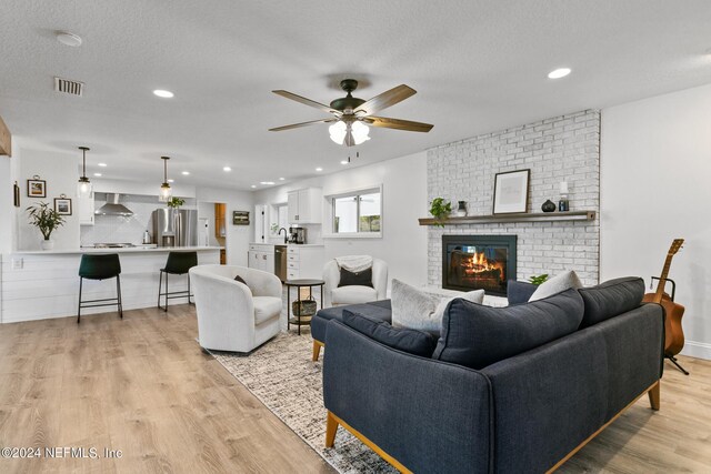 living room featuring a brick fireplace, ceiling fan, light wood-type flooring, and a textured ceiling