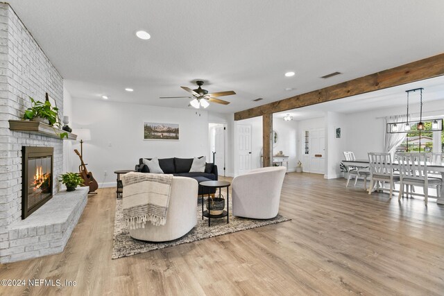 living room with a brick fireplace, beamed ceiling, a textured ceiling, light hardwood / wood-style flooring, and ceiling fan