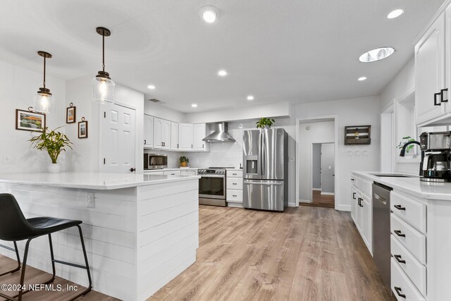 kitchen with light wood-type flooring, white cabinets, kitchen peninsula, wall chimney range hood, and appliances with stainless steel finishes