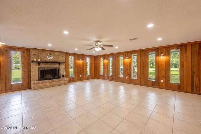 unfurnished living room with ceiling fan, wood walls, light tile patterned floors, a brick fireplace, and a textured ceiling