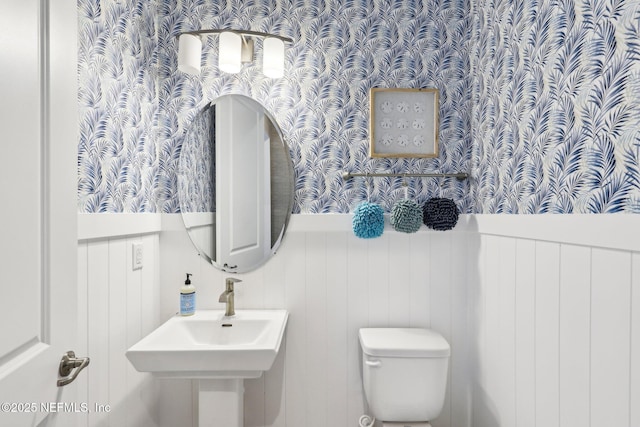 half bathroom with wainscoting, a sink, toilet, and wallpapered walls